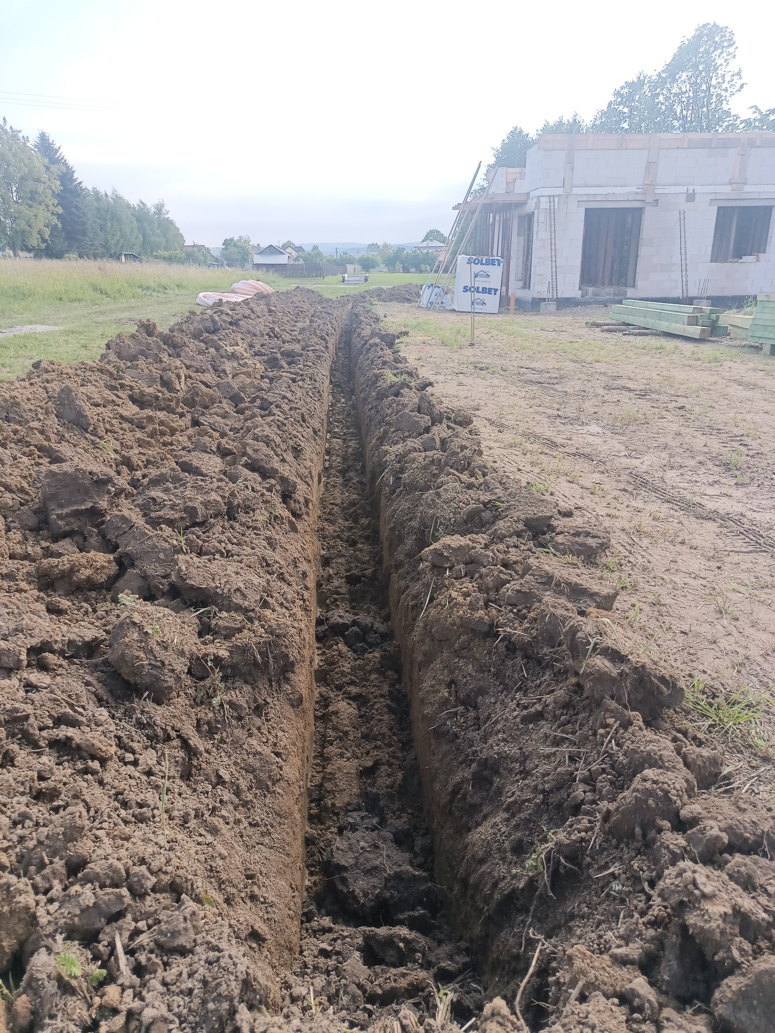 Wąski wykop w ziemi, prawdopodobnie pod instalację, z widokiem na budynek w trakcie budowy i materiały budowlane w tle. Ujęcie z poziomu gruntu.
