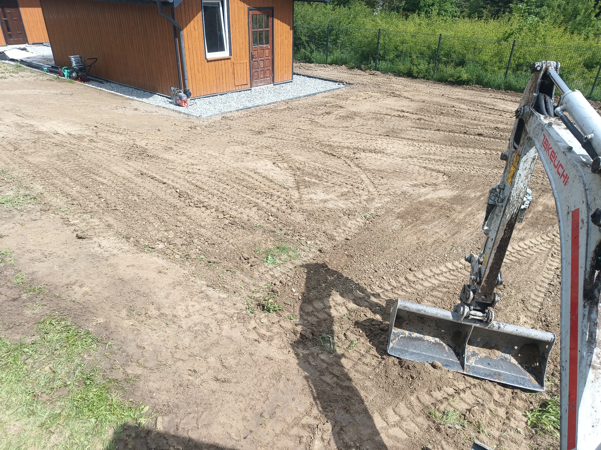 Mini koparka Takeuchi na placu budowy, wyrównany teren, widoczne ślady opon, drewniany budynek w tle. Przygotowanie terenu pod budowę.