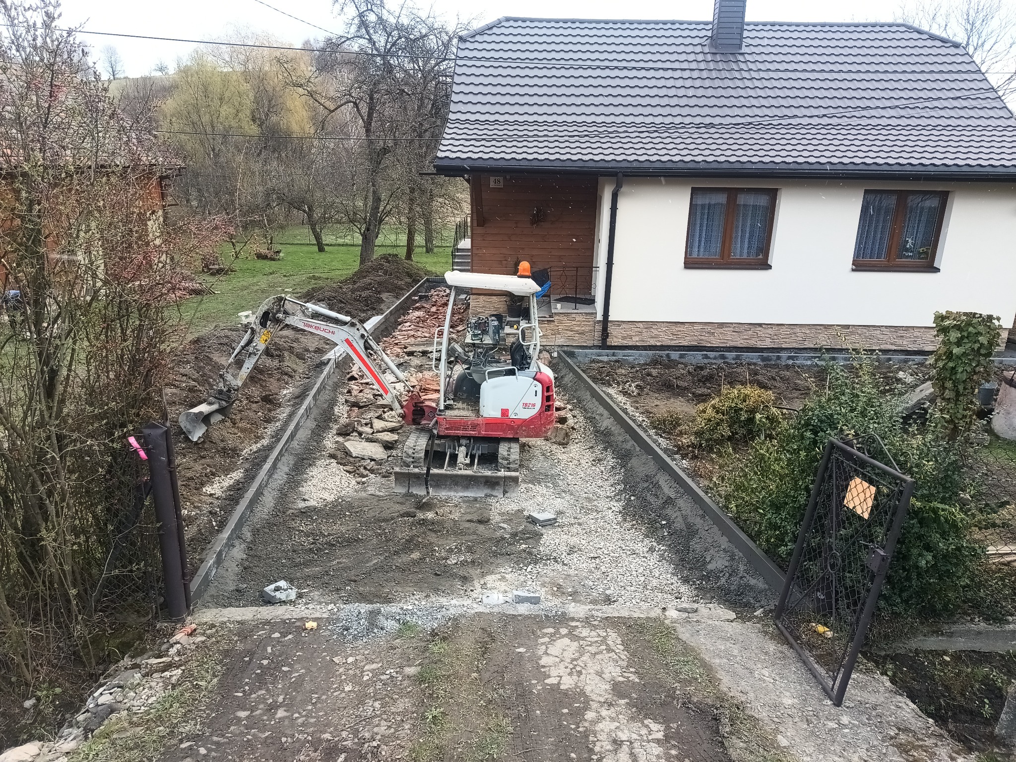 Mini koparka na placu budowy, przygotowuje podłoże z gruzu i kamieni pod chodnik lub podjazd. Widoczne obrzeża i dom w tle. Prace ziemne w toku.
