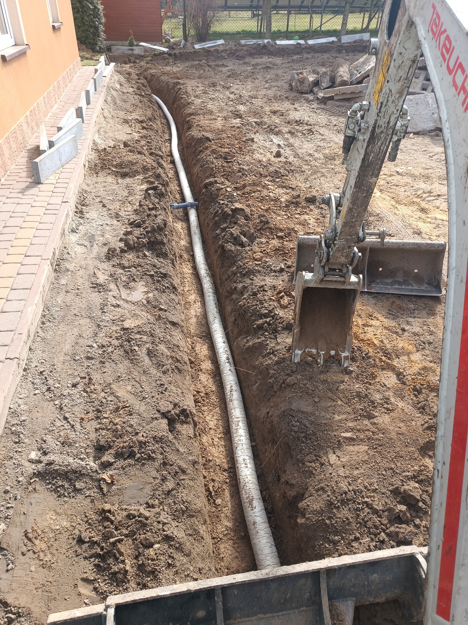 Wkopana rura w ziemi obok budynku, widoczna koparka. Etap prac ziemnych z użyciem ciężkiego sprzętu. Na pierwszym planie fragment koparki i wykopana ziemia.