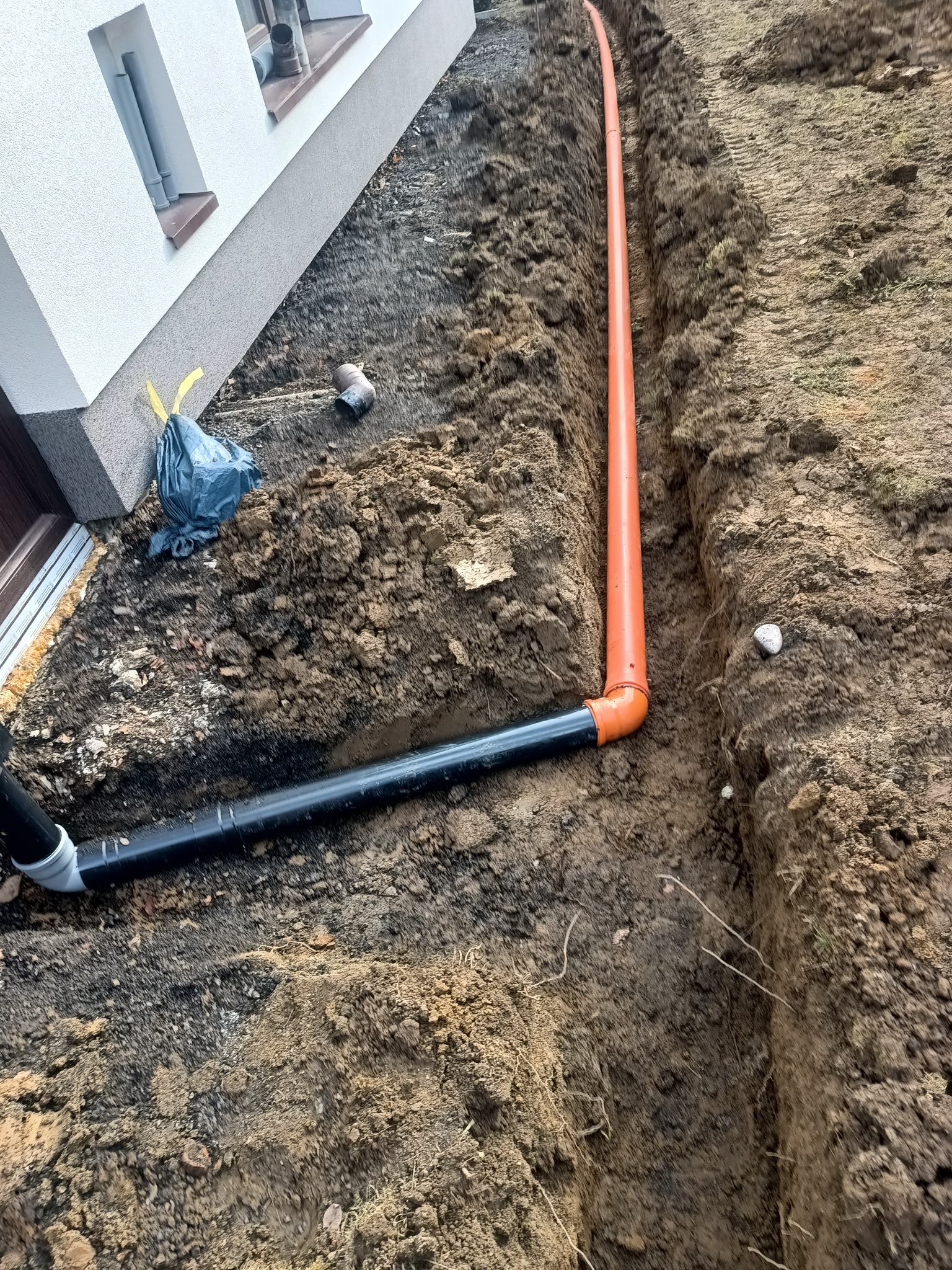 Wykop z ułożonymi rurami (pomarańczowa i czarna) przy fundamencie budynku. Widoczny fragment elewacji i okna. Grunt ziemny, przygotowany pod dalsze prace.