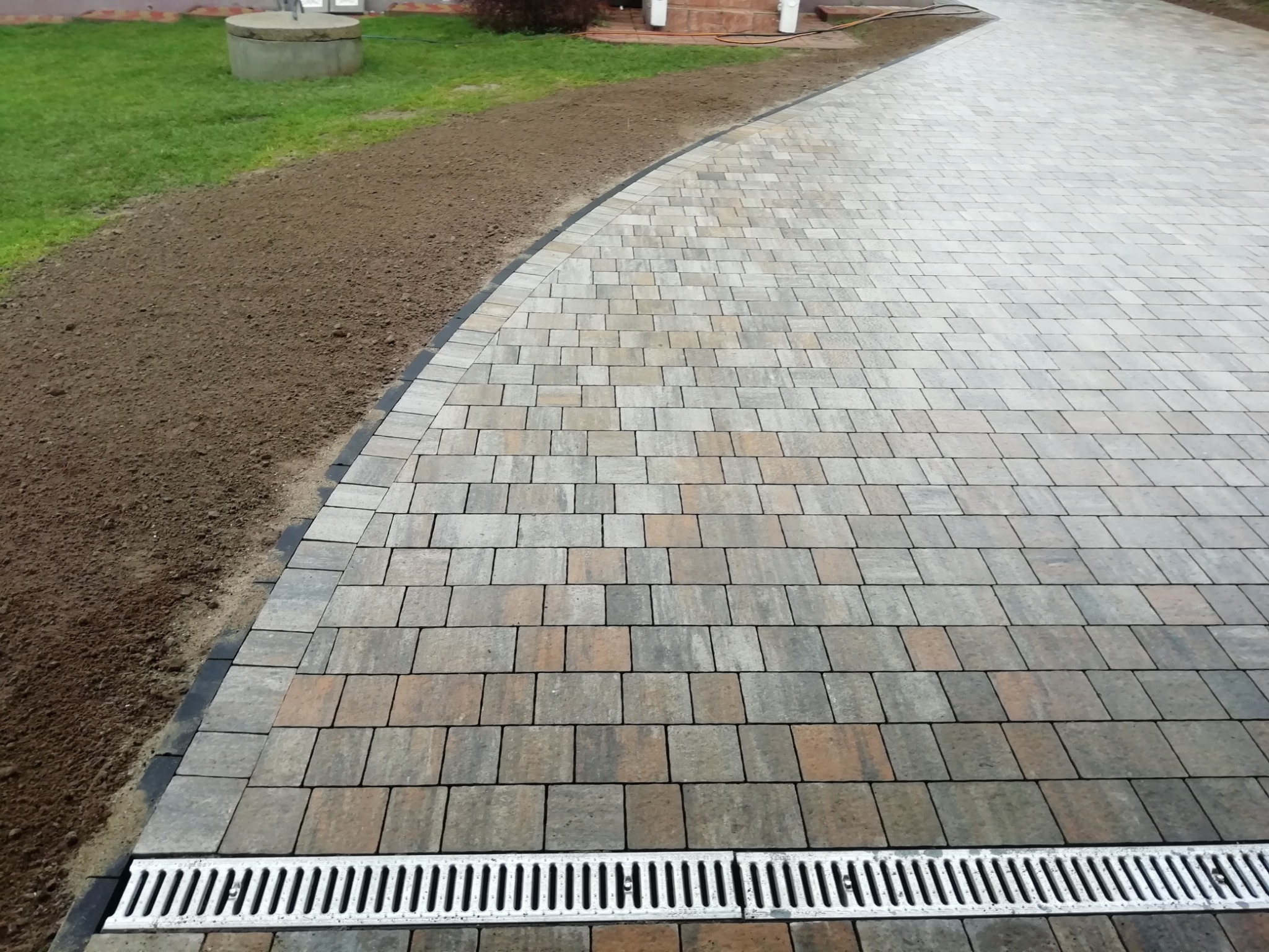 Ułożona kostka brukowa w odcieniach szarości i brązu, z widocznym odpływem liniowym i przygotowanym terenem pod trawnik. Nowoczesne i estetyczne wykończenie podjazdu.