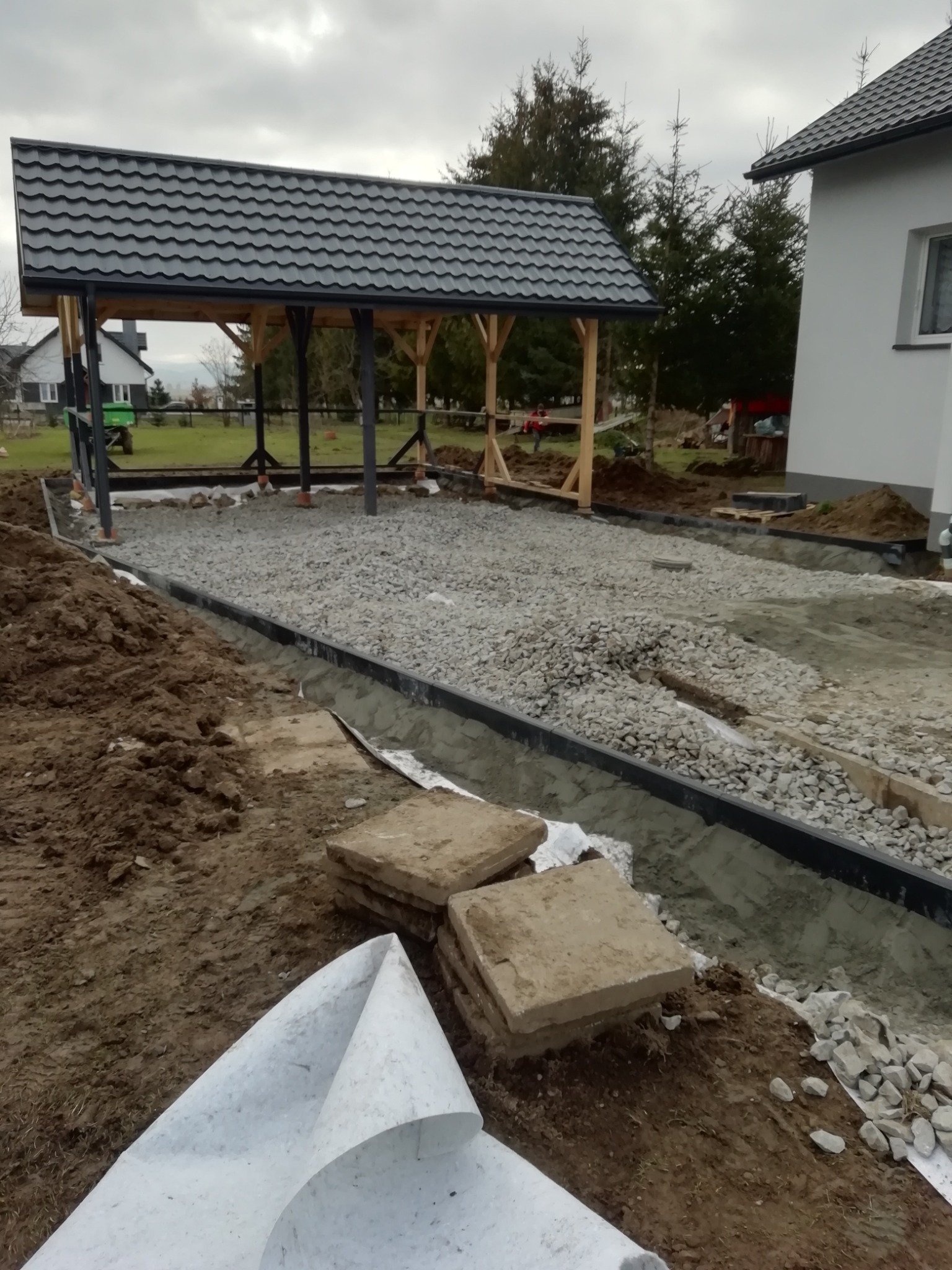 Przygotowanie podłoża pod kostkę brukową: podsypka z kamienia, obrzeża z tworzywa, geowłóknina. W tle wiata ogrodowa w trakcie budowy, dom i zieleń.