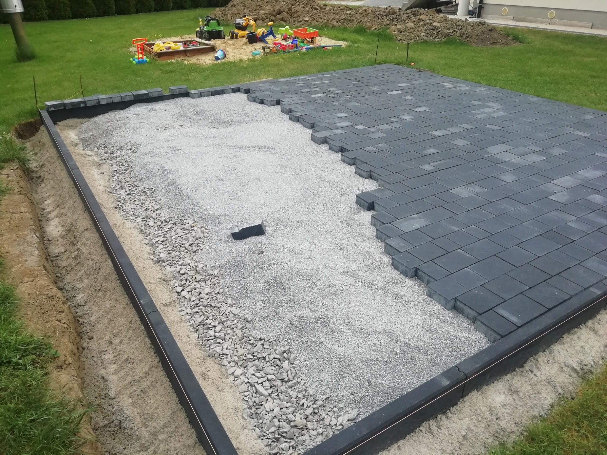 Częściowo ułożona, szara kostka brukowa na podsypce z kruszywa. Widoczne obrzeża i przygotowane podłoże. W tle trawnik i zabawki dziecięce.