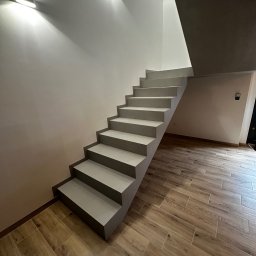 HUBERT HERDZIK WYKOŃCZENIA - Nowoczesne, minimalistyczne schody w odcieniach szarości, oświetlone punktowymi lampami, z drewnopodobną podłogą. Ujęcie z dołu, podkreślające geometrię.