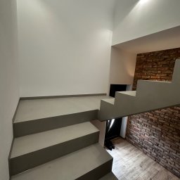 HUBERT HERDZIK WYKOŃCZENIA - Nowoczesne schody z szarego betonu prowadzą w górę, obok ściany z cegły i drewnianej podłogi. Minimalistyczny design, oświetlenie punktowe.