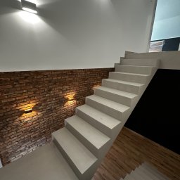 HUBERT HERDZIK WYKOŃCZENIA - Nowoczesne schody z oświetleniem ściennym, ceglana ściana, białe stopnie, drewnopodobna podłoga. Minimalistyczny design, ciepłe światło.