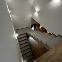 HUBERT HERDZIK WYKOŃCZENIA - Nowoczesne schody z podświetleniem LED na ścianach, wykończone drewnopodobnymi płytkami i cegłą. Widok z góry na jasne, minimalistyczne wnętrze.