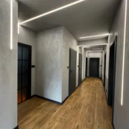 Wnętrze korytarza z drewnianą podłogą, szarymi ścianami o fakturze betonu i nowoczesnym oświetleniem LED wzdłuż sufitu i ścian. Czarne drzwi.