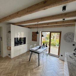 HUBERT HERDZIK WYKOŃCZENIA - Jasna kuchnia z parkietem w jodełkę, drewnianymi belkami stropowymi i przeszklonym wyjściem na ogród. Nowoczesne, białe meble kuchenne.