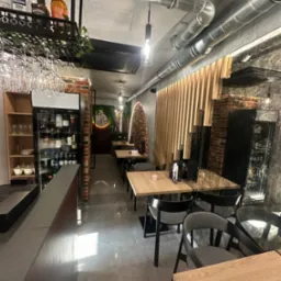 Wnętrze restauracji z surowym wykończeniem: odsłonięte rury wentylacyjne, cegła, drewniane lamele. Stoły z szarymi krzesłami, chłodziarka z napojami, dekoracje roślinne.