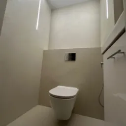 Minimalistyczna toaleta w odcieniach beżu z podtynkową spłuczką i liniowym oświetleniem LED, tworząca nowoczesny i elegancki wystrój wnętrza.