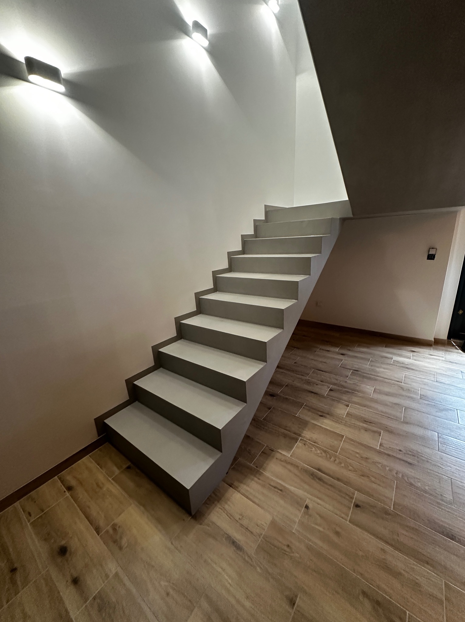 Nowoczesne, minimalistyczne schody w odcieniach szarości, oświetlone punktowymi lampami, z drewnopodobną podłogą. Ujęcie z dołu, podkreślające geometrię.