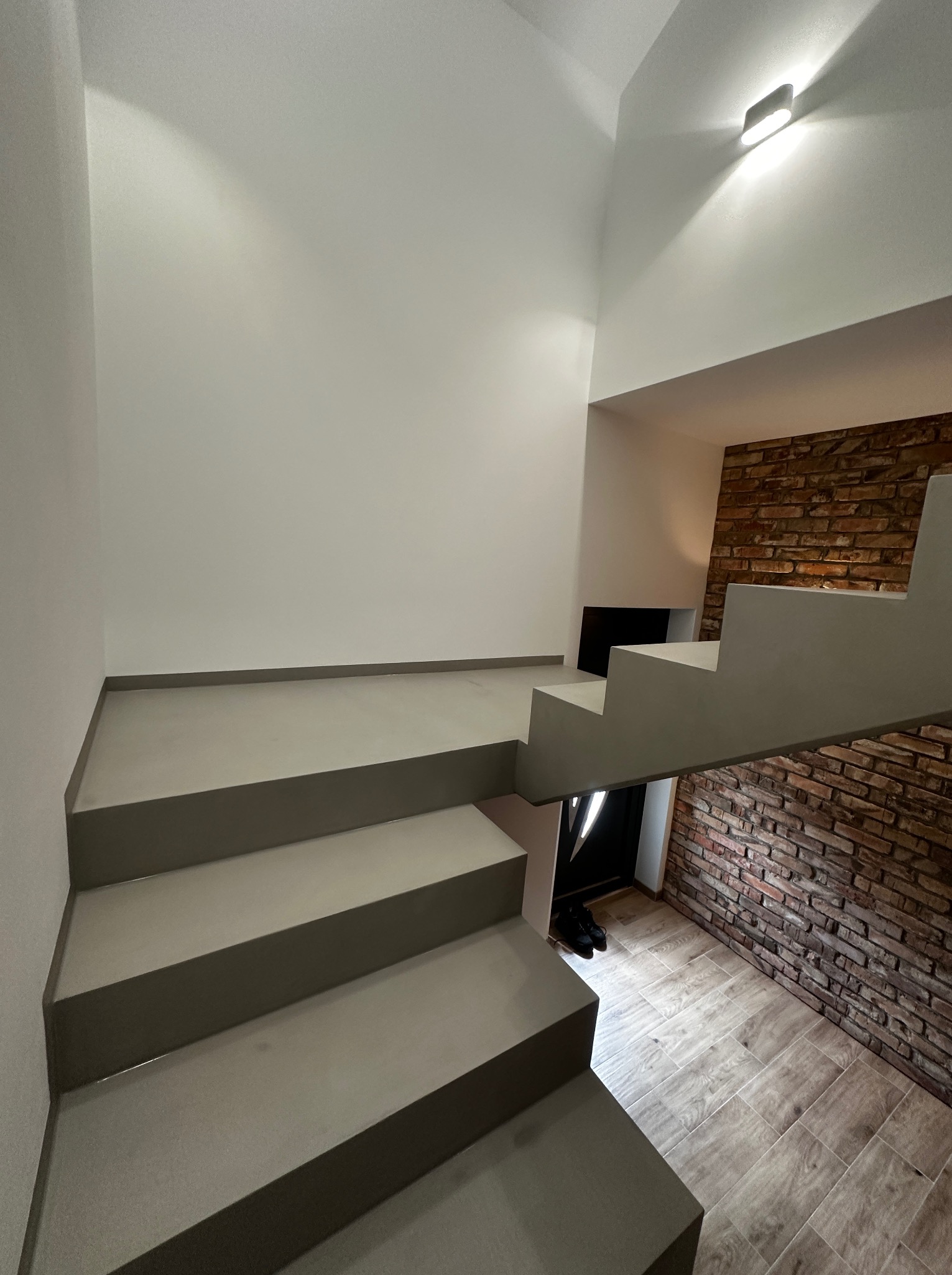 Nowoczesne schody z szarego betonu prowadzą w górę, obok ściany z cegły i drewnianej podłogi. Minimalistyczny design, oświetlenie punktowe.