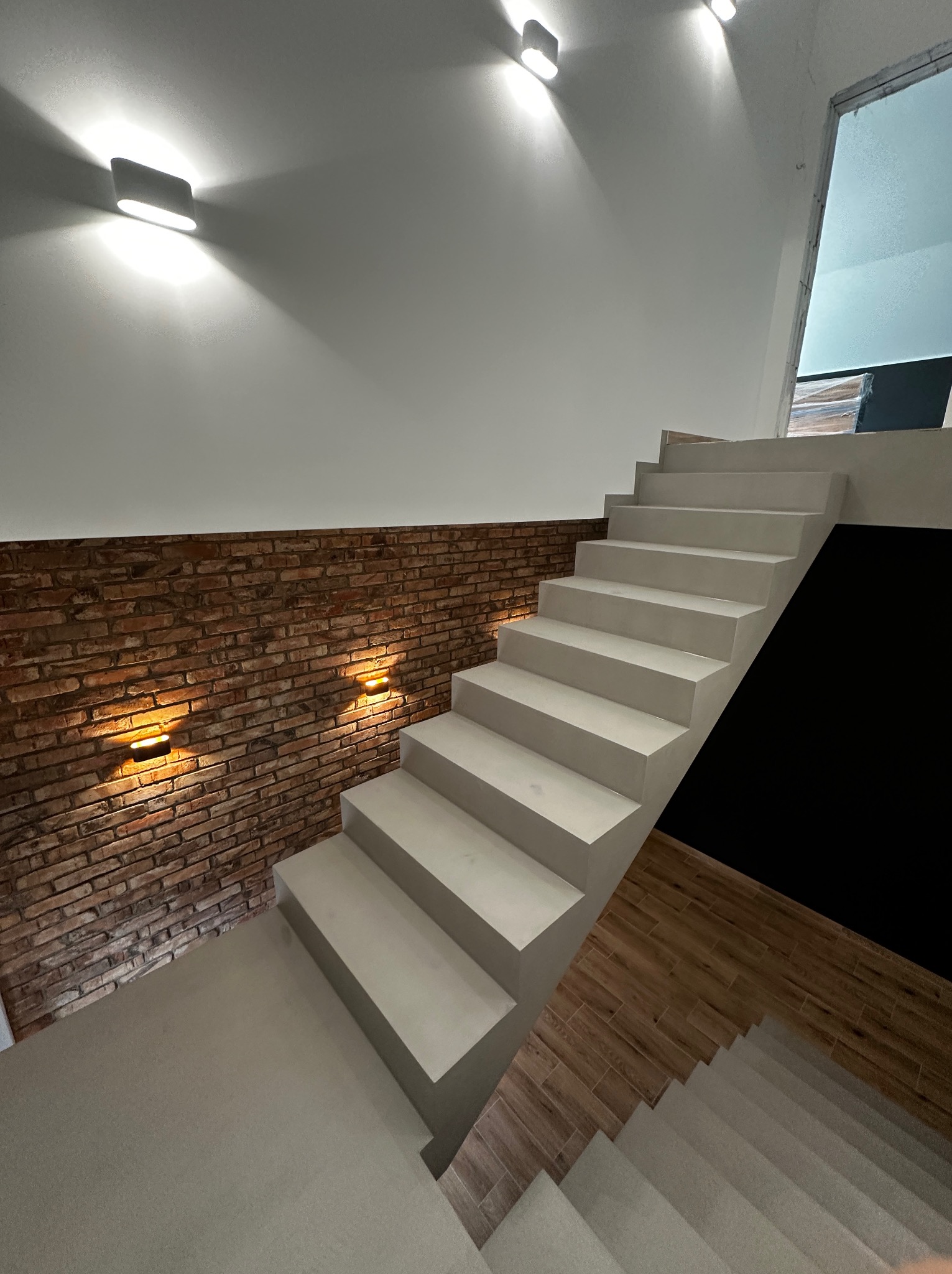 Nowoczesne schody z oświetleniem ściennym, ceglana ściana, białe stopnie, drewnopodobna podłoga. Minimalistyczny design, ciepłe światło.