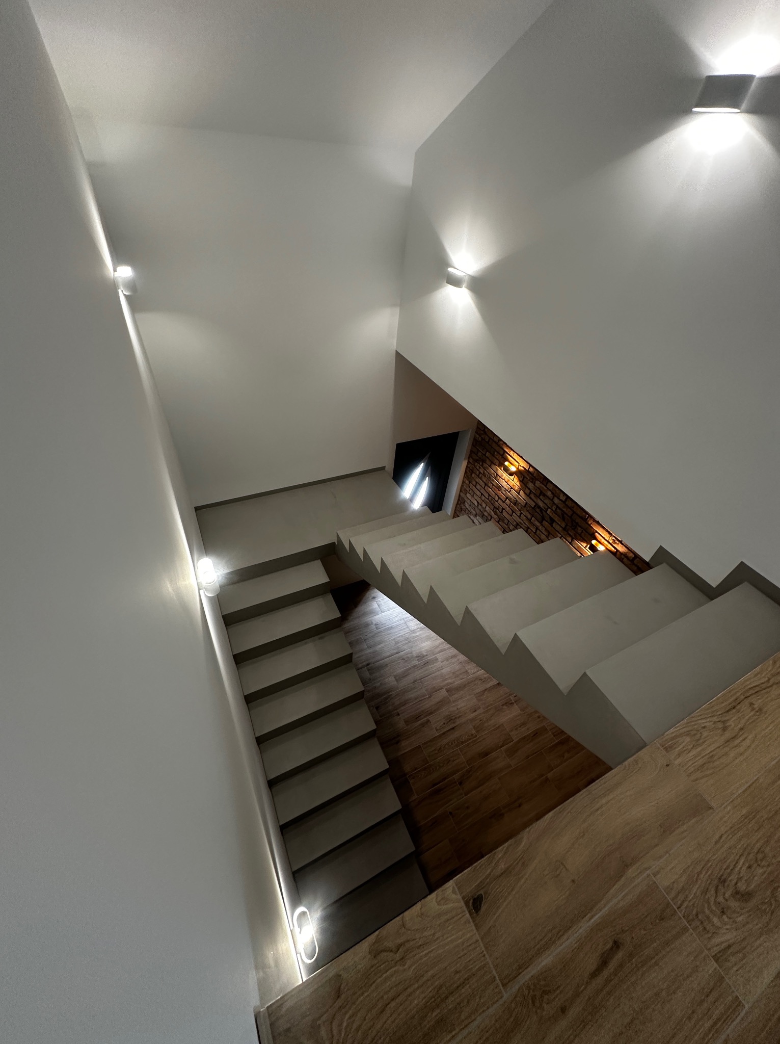 Nowoczesne schody z podświetleniem LED na ścianach, wykończone drewnopodobnymi płytkami i cegłą. Widok z góry na jasne, minimalistyczne wnętrze.