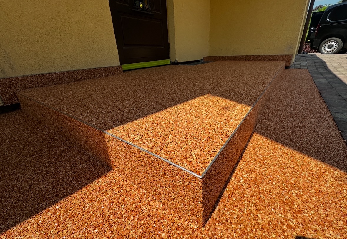 Wykończenie schodów wejściowych żywicą epoksydową z dekoracyjnym kruszywem kwarcowym w odcieniach brązu. Widoczne detale wykończeniowe i fragment elewacji budynku.