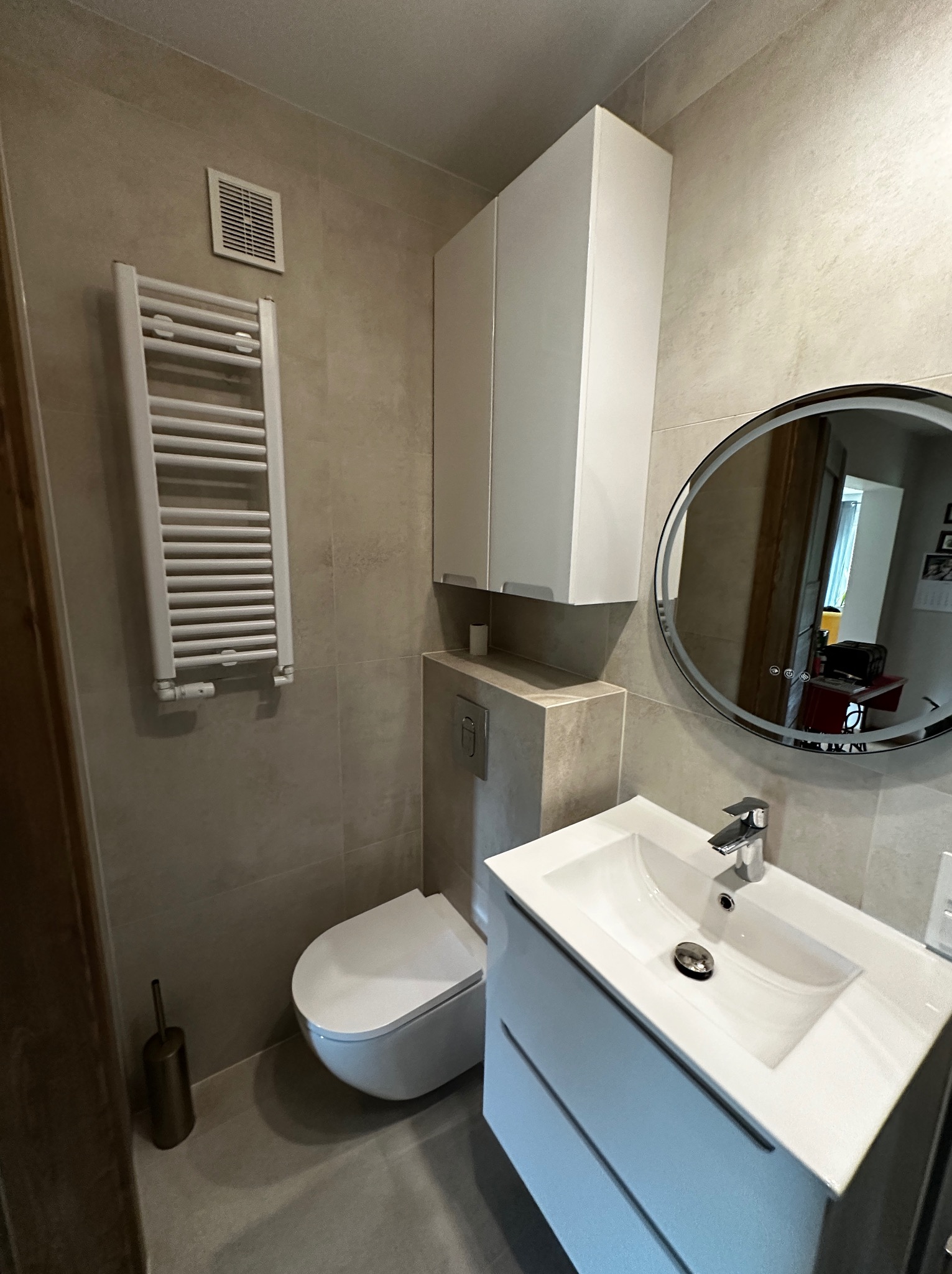 Jasna łazienka po remoncie: umywalka z szafką, toaleta, grzejnik drabinkowy i okrągłe lustro z podświetleniem. Minimalistyczny design, beżowe płytki.