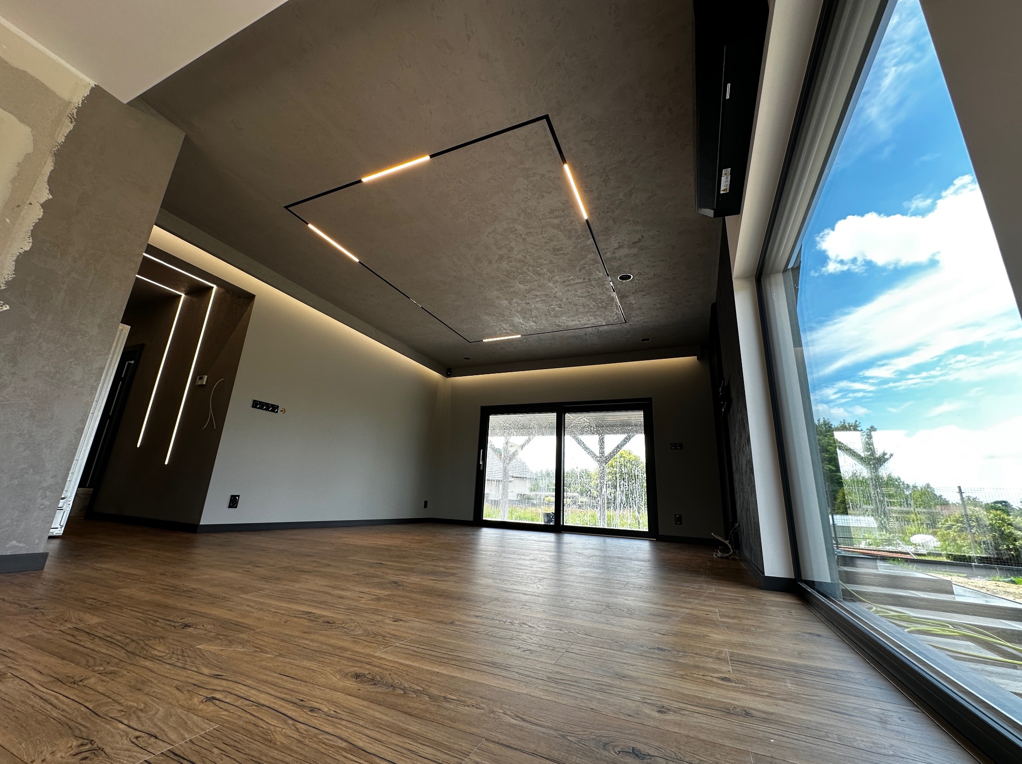 Nowoczesne wnętrze z drewnianą podłogą, betonowym sufitem z oświetleniem LED i dużym oknem panoramicznym z widokiem na zieleń. Częściowo wykończone ściany.