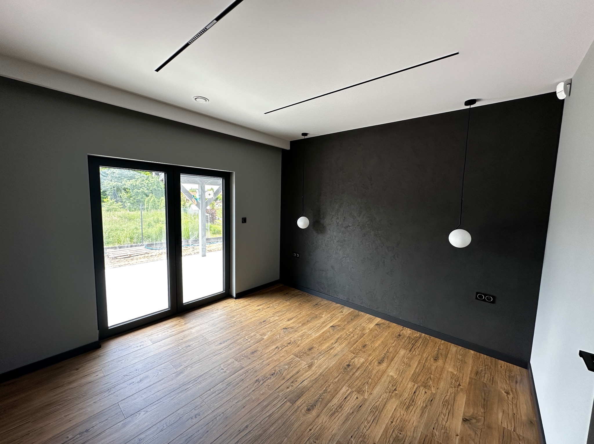 Nowoczesne wnętrze z drewnianą podłogą, czarną ścianą z fakturą i minimalistycznym oświetleniem. Duże okno balkonowe wpuszcza światło. Widok na zieleń za oknem.