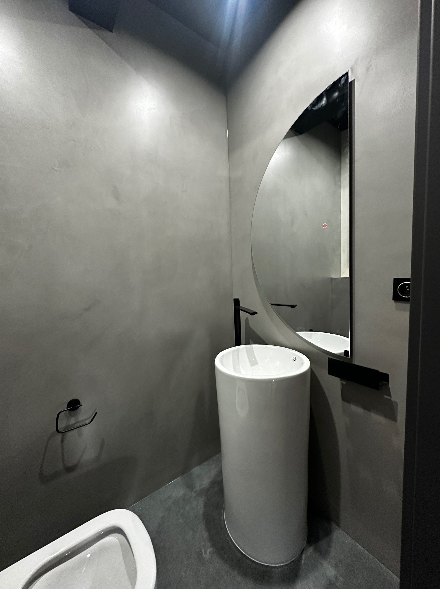 Minimalistyczna łazienka w odcieniach szarości z okrągłym lustrem i białą umywalką na postumencie. Ściany wykończone w stylu betonu architektonicznego.