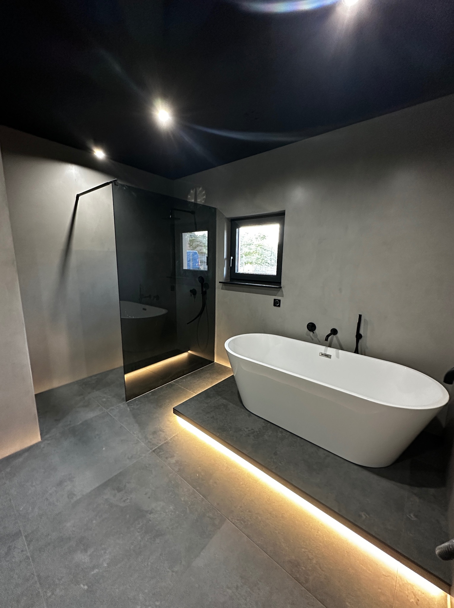 Nowoczesna łazienka w odcieniach szarości z czarnym sufitem, wolnostojącą białą wanną i przeszklonym prysznicem. Podświetlenie LED dodaje ciepła.