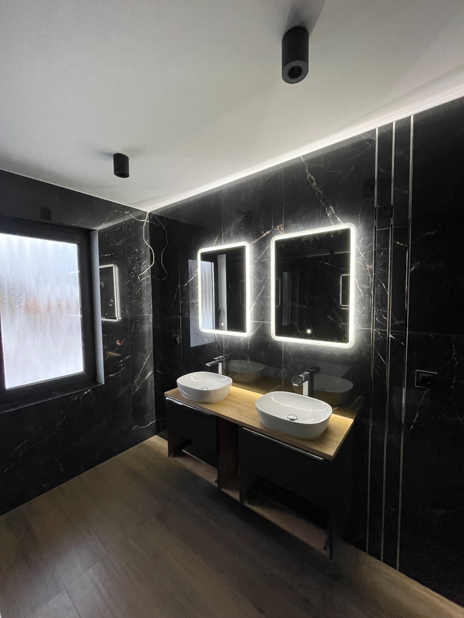 Nowoczesna łazienka z czarnymi marmurowymi płytkami, podświetlanymi lustrami i drewnianym blatem pod umywalki, oświetlenie punktowe.