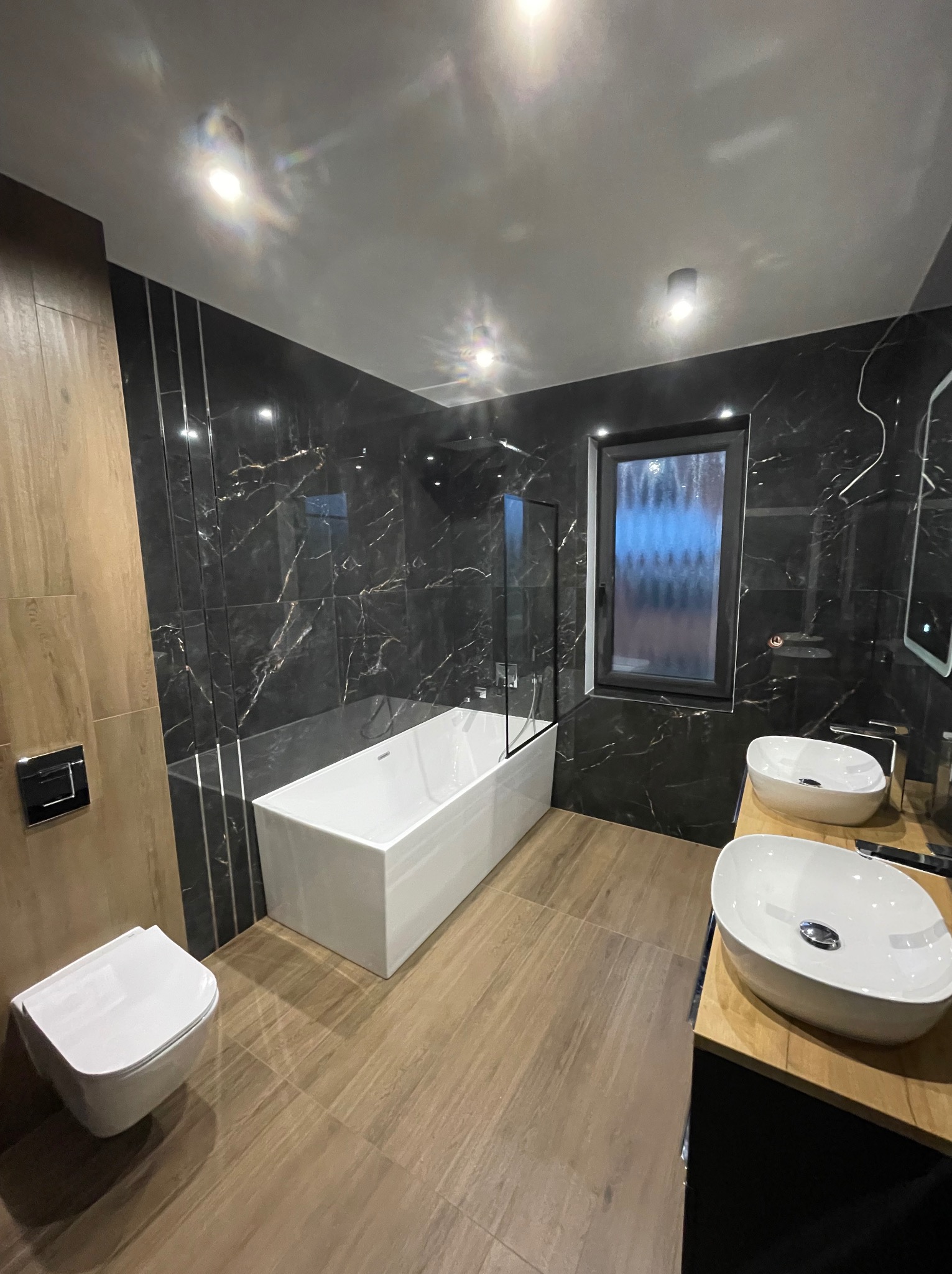 Nowoczesna łazienka z czarnymi marmurowymi płytkami, białą wanną i umywalkami nablatowymi. Drewnopodobna podłoga i oświetlenie punktowe.