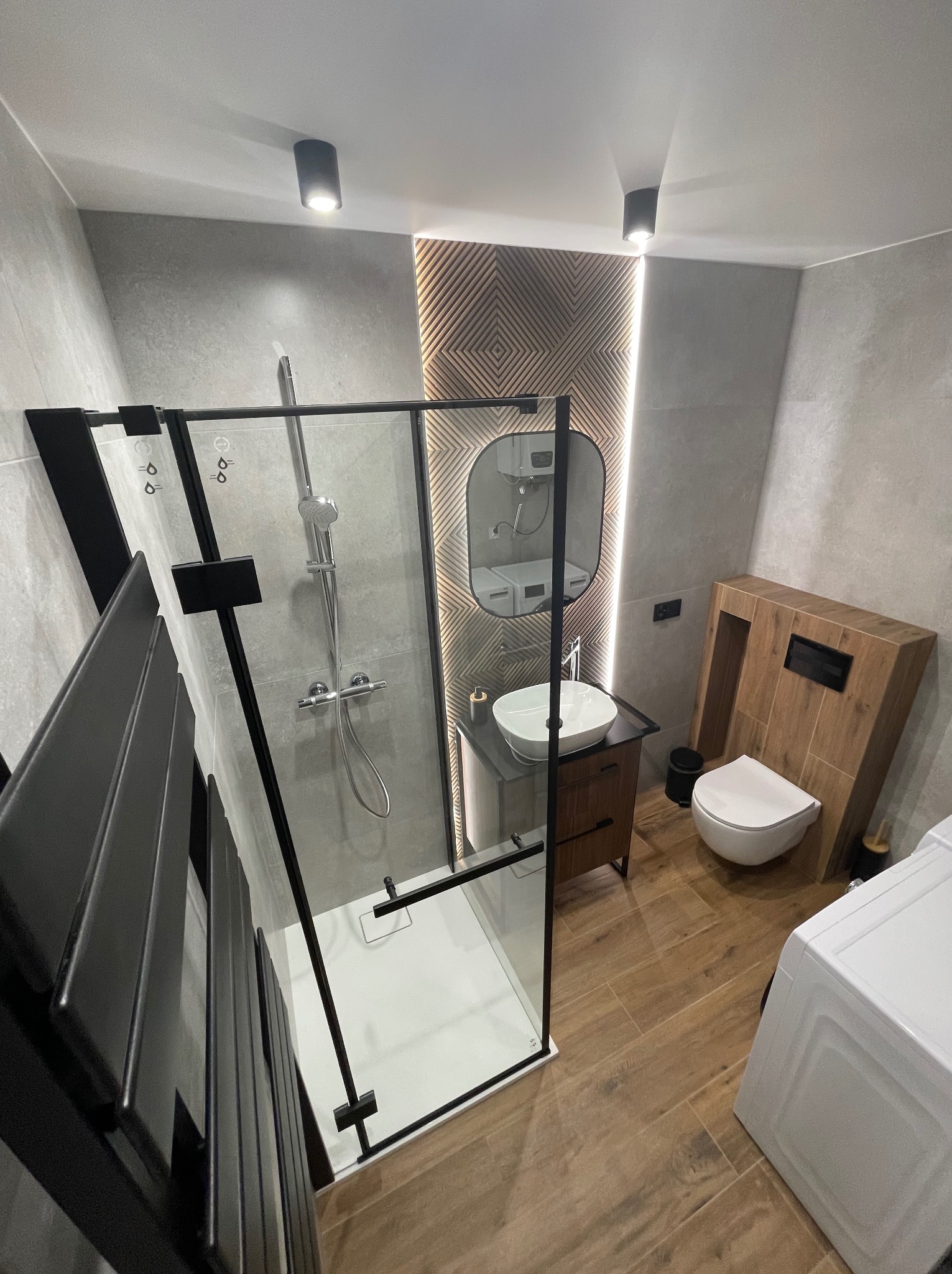 Nowoczesna łazienka z kabiną prysznicową typu walk-in z czarnymi ramami, drewnianą zabudową toalety i umywalki oraz oświetleniem punktowym.
