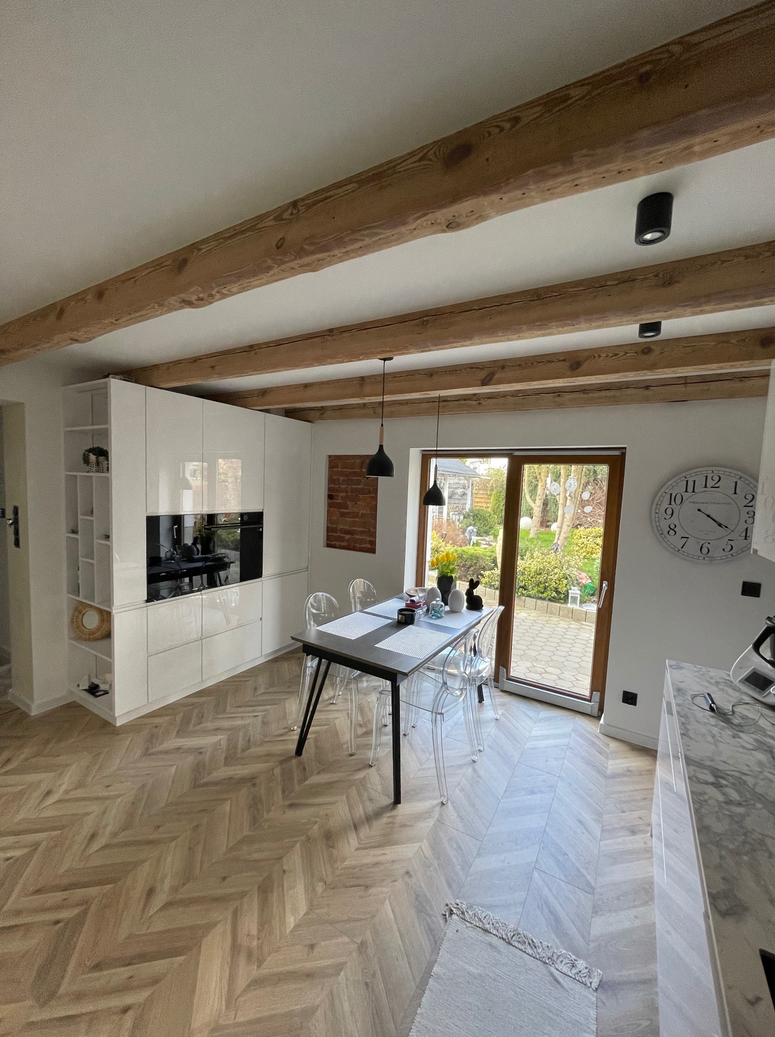 Jasna kuchnia z parkietem w jodełkę, drewnianymi belkami stropowymi i przeszklonym wyjściem na ogród. Nowoczesne, białe meble kuchenne.