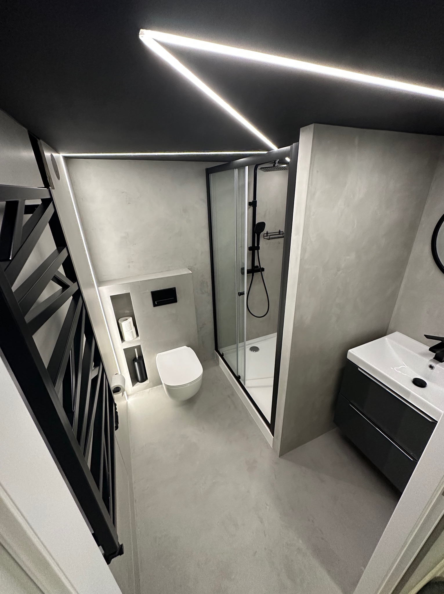 Wnętrze łazienki po remoncie: kabina prysznicowa, umywalka z szafką, toaleta podwieszana i grzejnik drabinkowy. Szare ściany, czarne akcenty i ledowe oświetlenie.