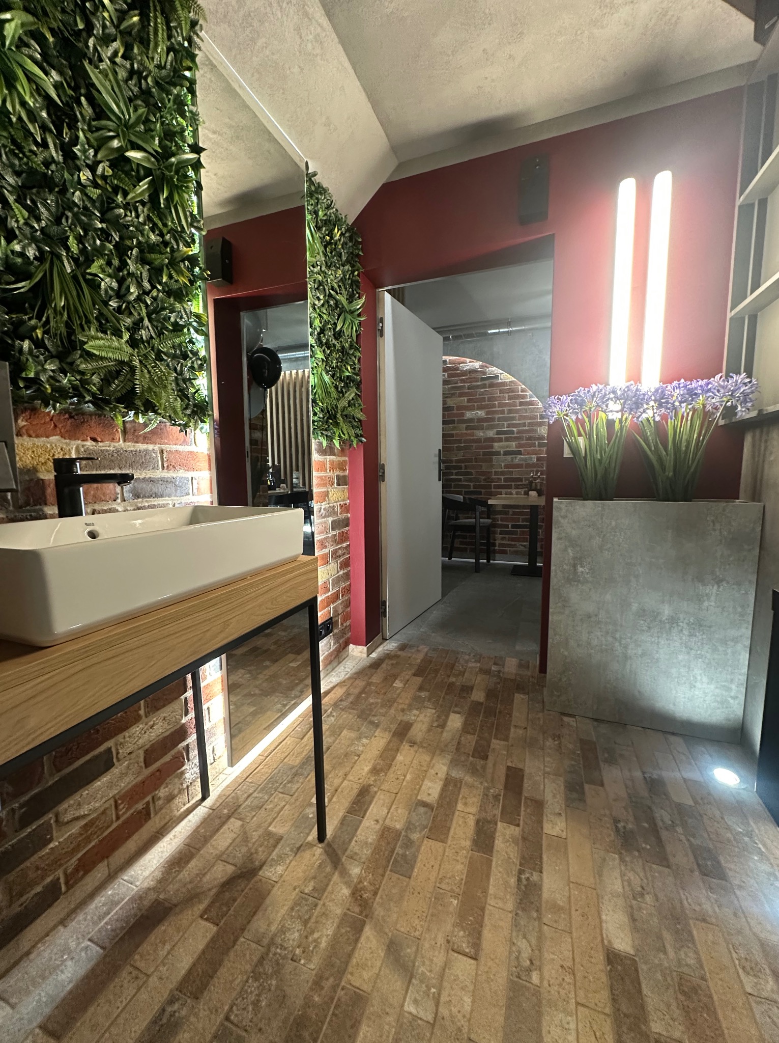 Nowoczesna łazienka z ceglaną ścianą, umywalką na drewnianym blacie i pionową zielenią. Widoczny fragment podłogi z cegły i oświetlenie.