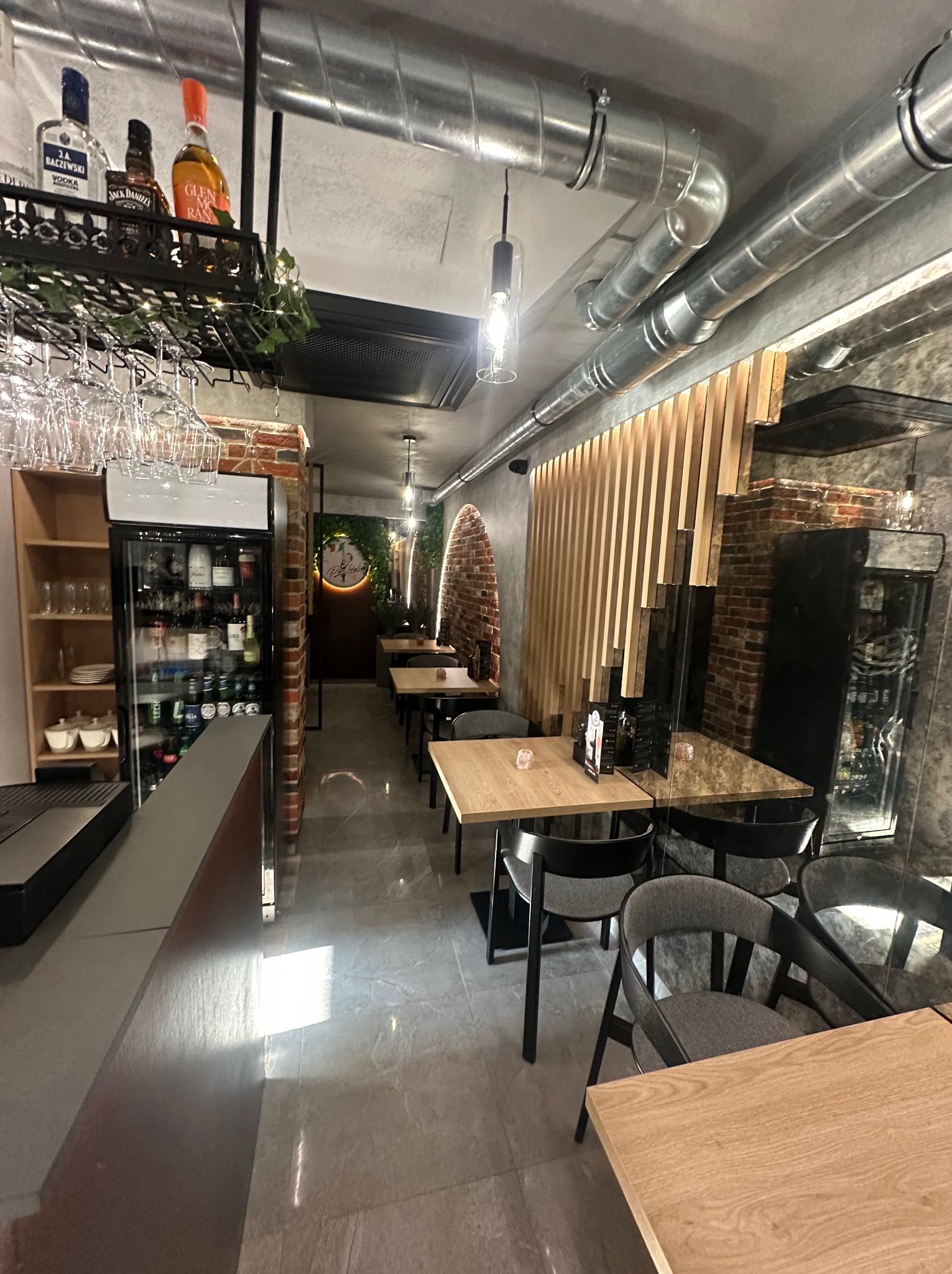 Wnętrze restauracji z surowym wykończeniem: odsłonięte rury wentylacyjne, cegła, drewniane lamele. Stoły z szarymi krzesłami, chłodziarka z napojami, dekoracje roślinne.