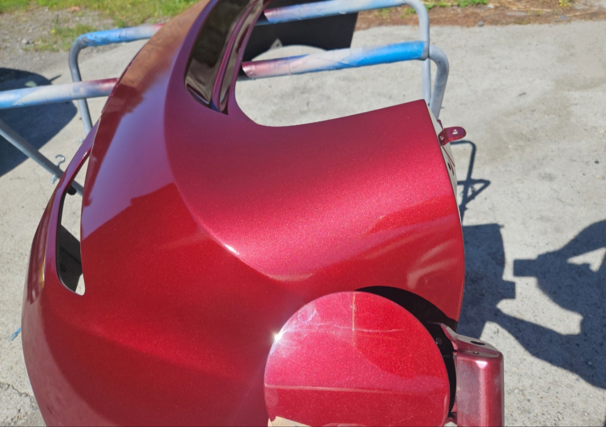 Błyszczący, bordowy zderzak samochodowy po lakierowaniu, zawieszony na stojaku. Widoczne drobne rysy na powierzchni, ostre światło słoneczne.