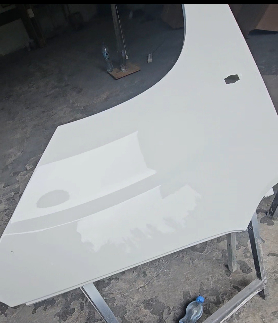 Biały błotnik samochodowy po lakierowaniu, leżący na metalowym stojaku. Widoczne odbicia światła na gładkiej powierzchni lakieru. Detal z otworem po prawej stronie.