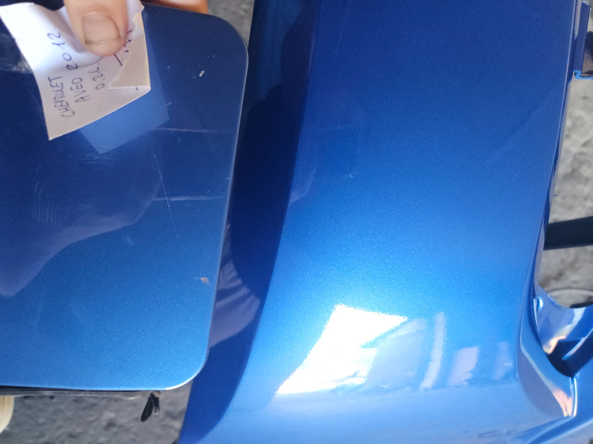Błękitny błotnik Chevrolet Aveo 2012 z rysami, w trakcie doboru lakieru. Notatka z danymi odcienia trzymana w dłoni, zbliżenie na uszkodzoną powierzchnię.