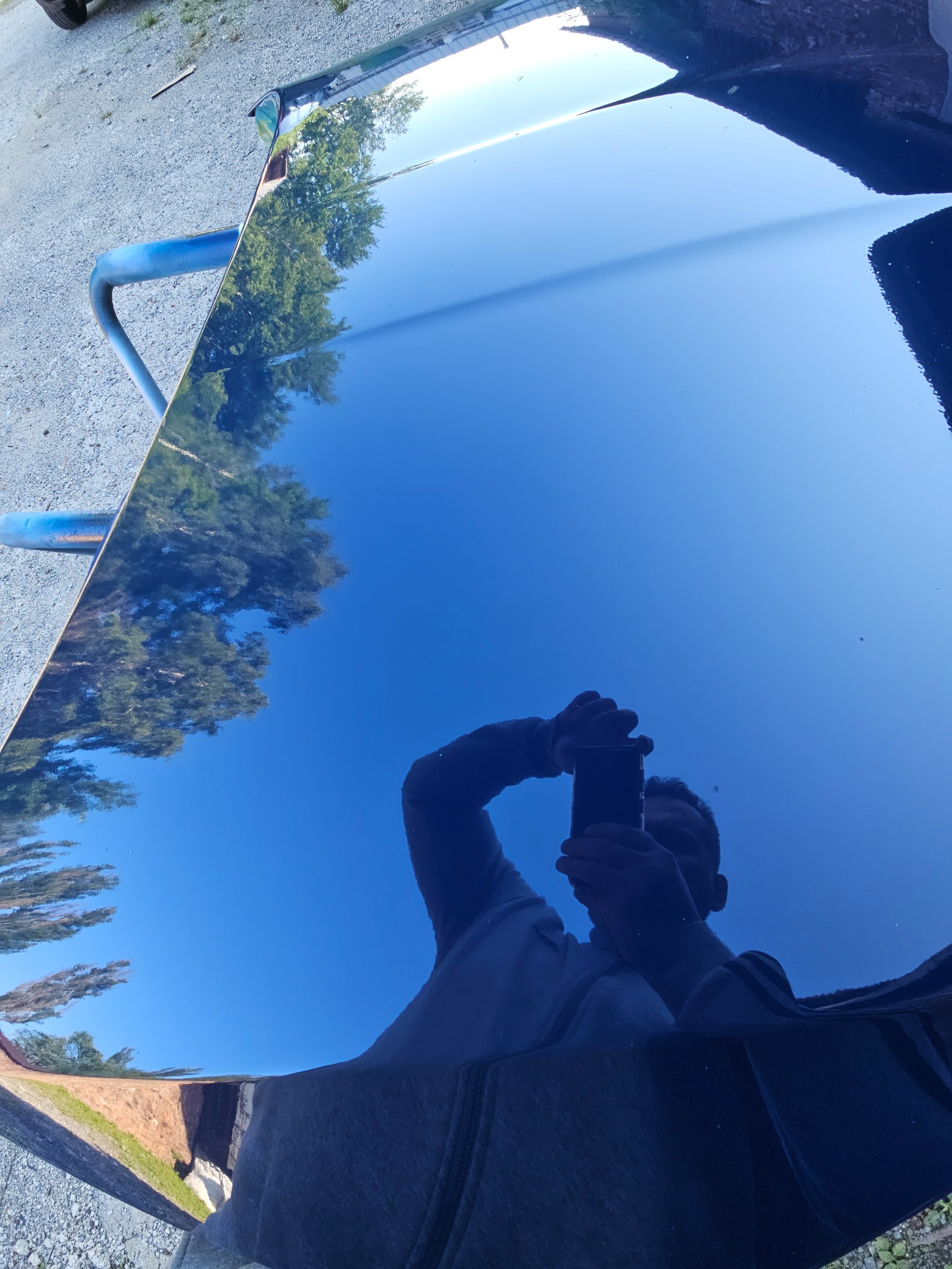 Błyszcząca, granatowa maska samochodu po polerowaniu odbija niebo i drzewa; widoczne odbicie fotografa z telefonem w dłoni. Auto na stojaku.