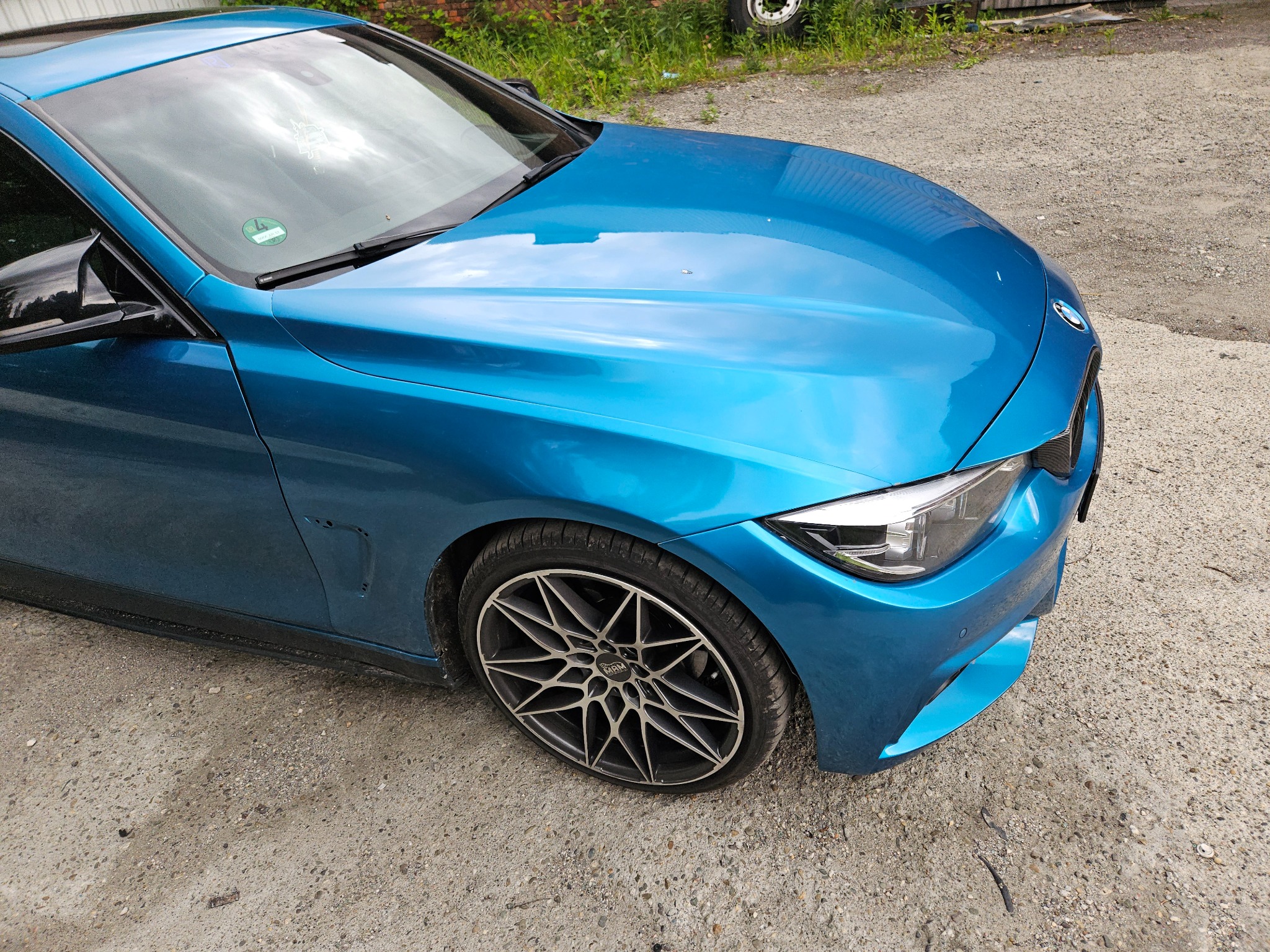 Błękitny samochód BMW z felgami aluminiowymi na żwirowym podłożu. Widoczny fragment karoserii z reflektorem i kołem.