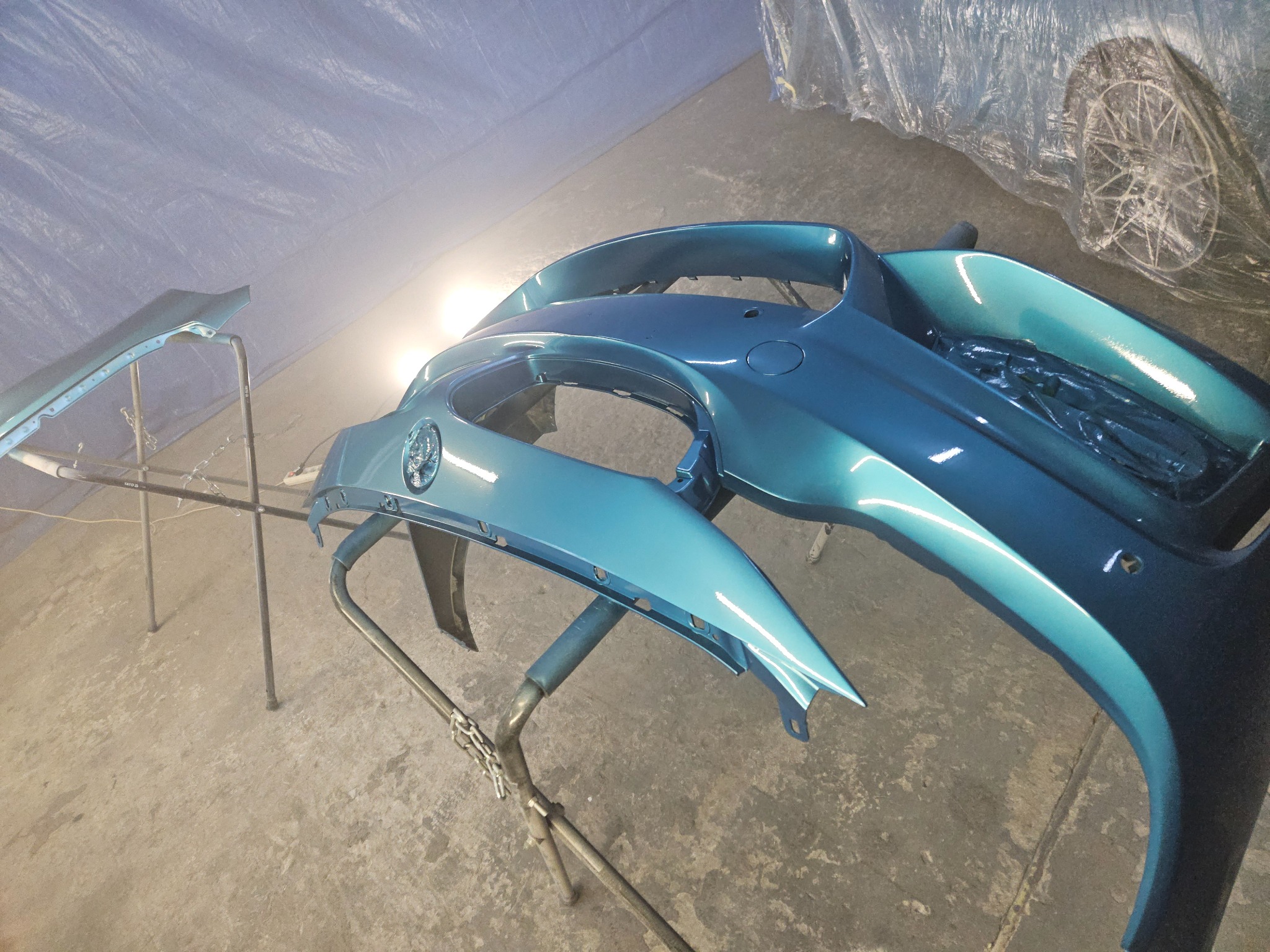 Turkusowy, świeżo polakierowany zderzak samochodowy na stojaku warsztatowym, z widocznym odbiciem światła, w tle fragment koła i folia ochronna.