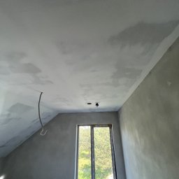 Zabudowy Wnętrz Paweł Wójcik - Poddasze po szpachlowaniu, widoczne niedoskonałości faktury na ścianach i suficie. Okno z widokiem na zieleń. Wystający kabel elektryczny, surowy stan wykończenia.