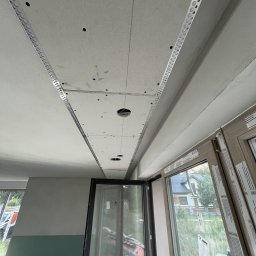 Zabudowy Wnętrz Paweł Wójcik - Sufit z zamontowaną płytą karton-gips, widoczne profile i otwory na oświetlenie, zarysowany czerwoną linią. W tle okno z widokiem na dom.