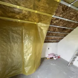 Skośne poddasze w trakcie prac: widoczna izolacja wełną mineralną, metalowy stelaż i folia ochronna. W tle drabina i resztki materiałów budowlanych.