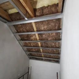 Metalowy stelaż sufitu podwieszanego z izolacją z wełny mineralnej, widok z dołu, przygotowanie do montażu płyt, ściany pomalowane na biało.