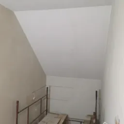 Poddasze w trakcie prac wykończeniowych: szpachlowana ściana w odcieniach beżu, rusztowanie z deskami i widoczna puszka elektryczna. Skos dachu.