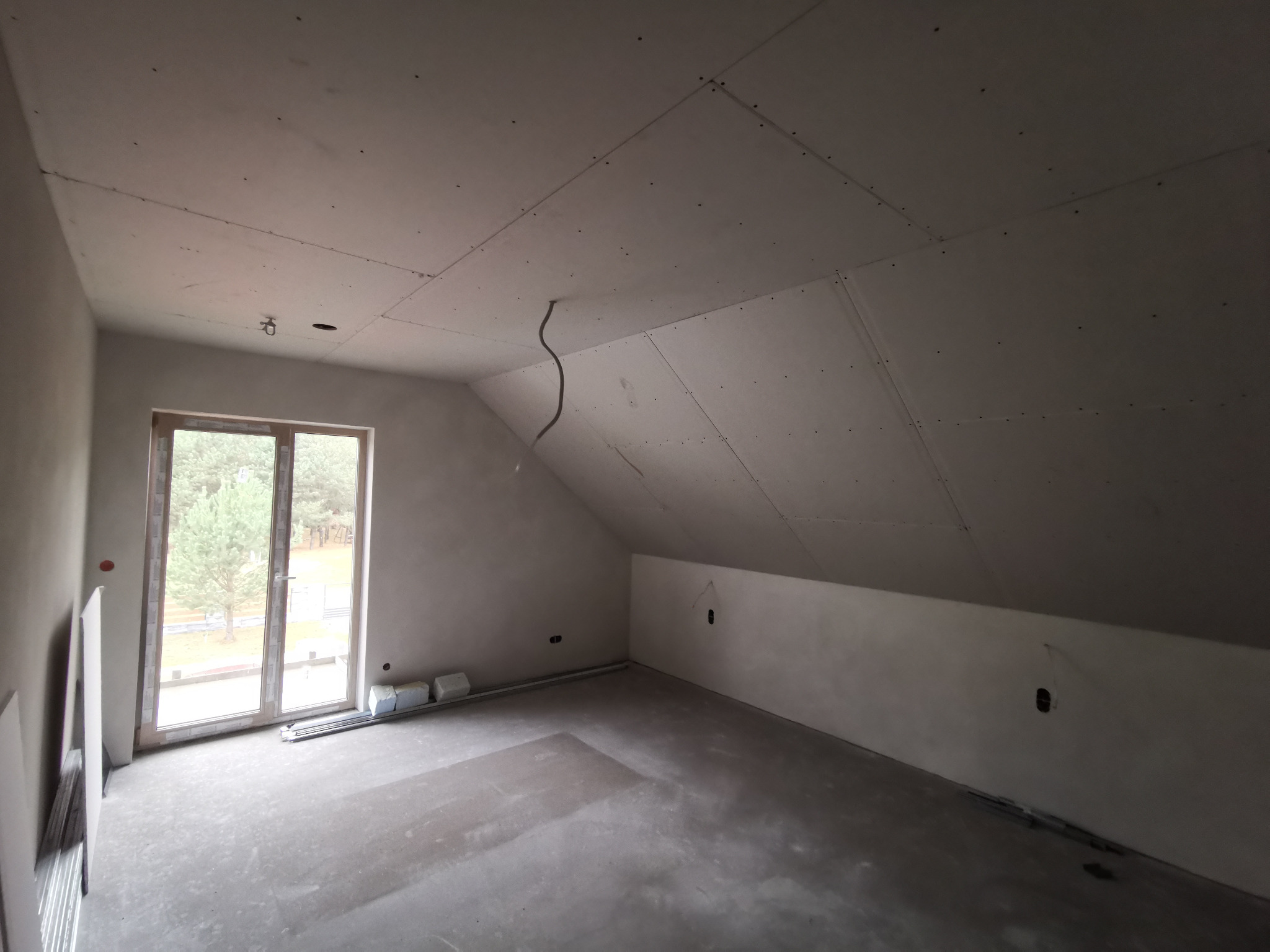 Poddasze po montażu płyt gipsowych na skosach i suficie, widoczne okno balkonowe i przygotowana instalacja elektryczna. Surowy stan, szare ściany, betonowa podłoga.
