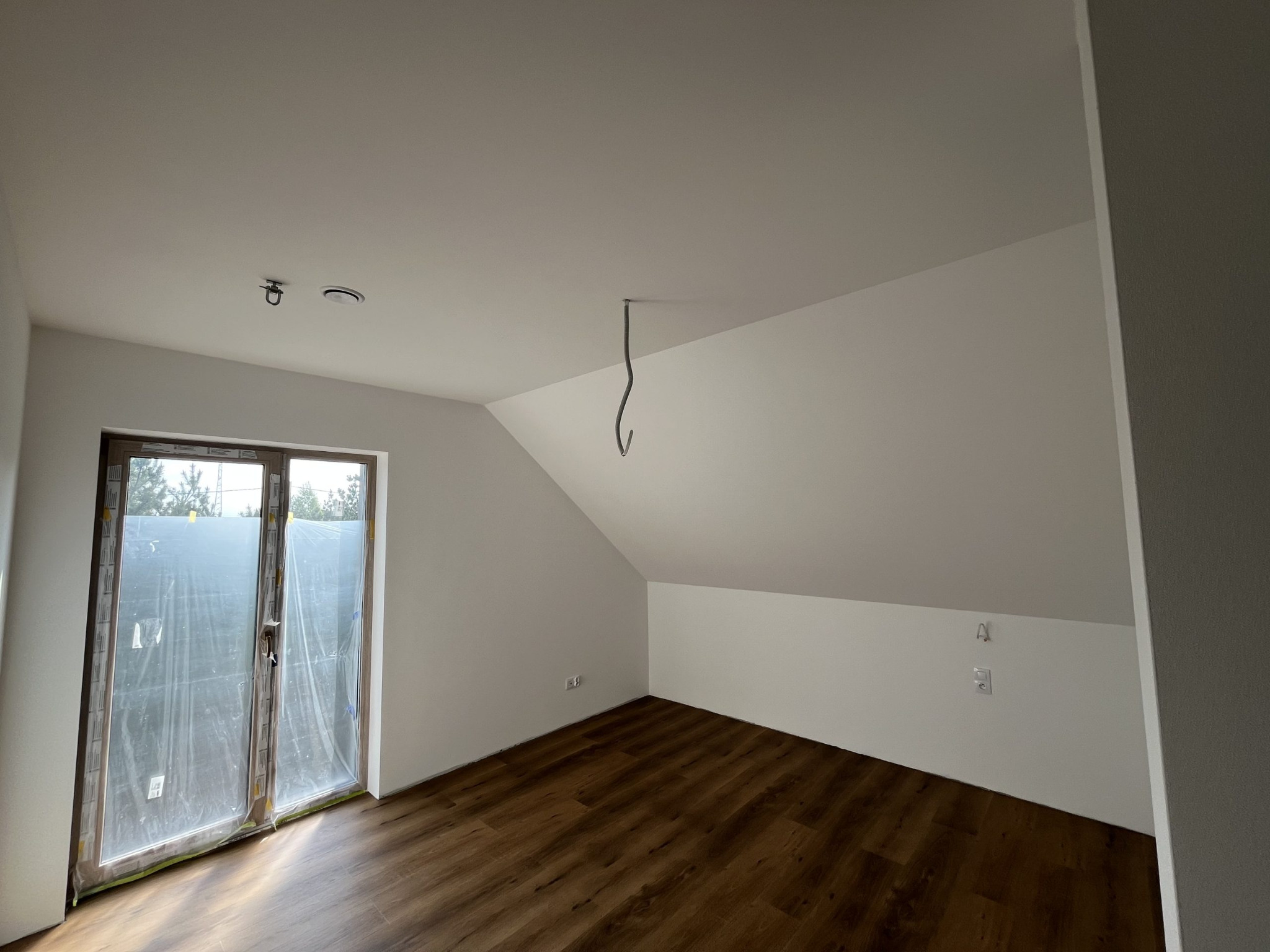 Poddasze po remoncie: białe ściany, brązowe panele, okno zabezpieczone folią. Widoczne gniazdka i wystający kabel ze sufitu. Skos dachu nadaje pomieszczeniu charakteru.