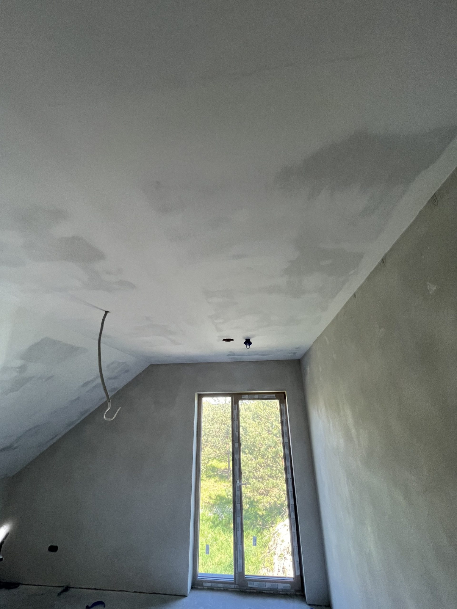 Poddasze po szpachlowaniu, widoczne niedoskonałości faktury na ścianach i suficie. Okno z widokiem na zieleń. Wystający kabel elektryczny, surowy stan wykończenia.