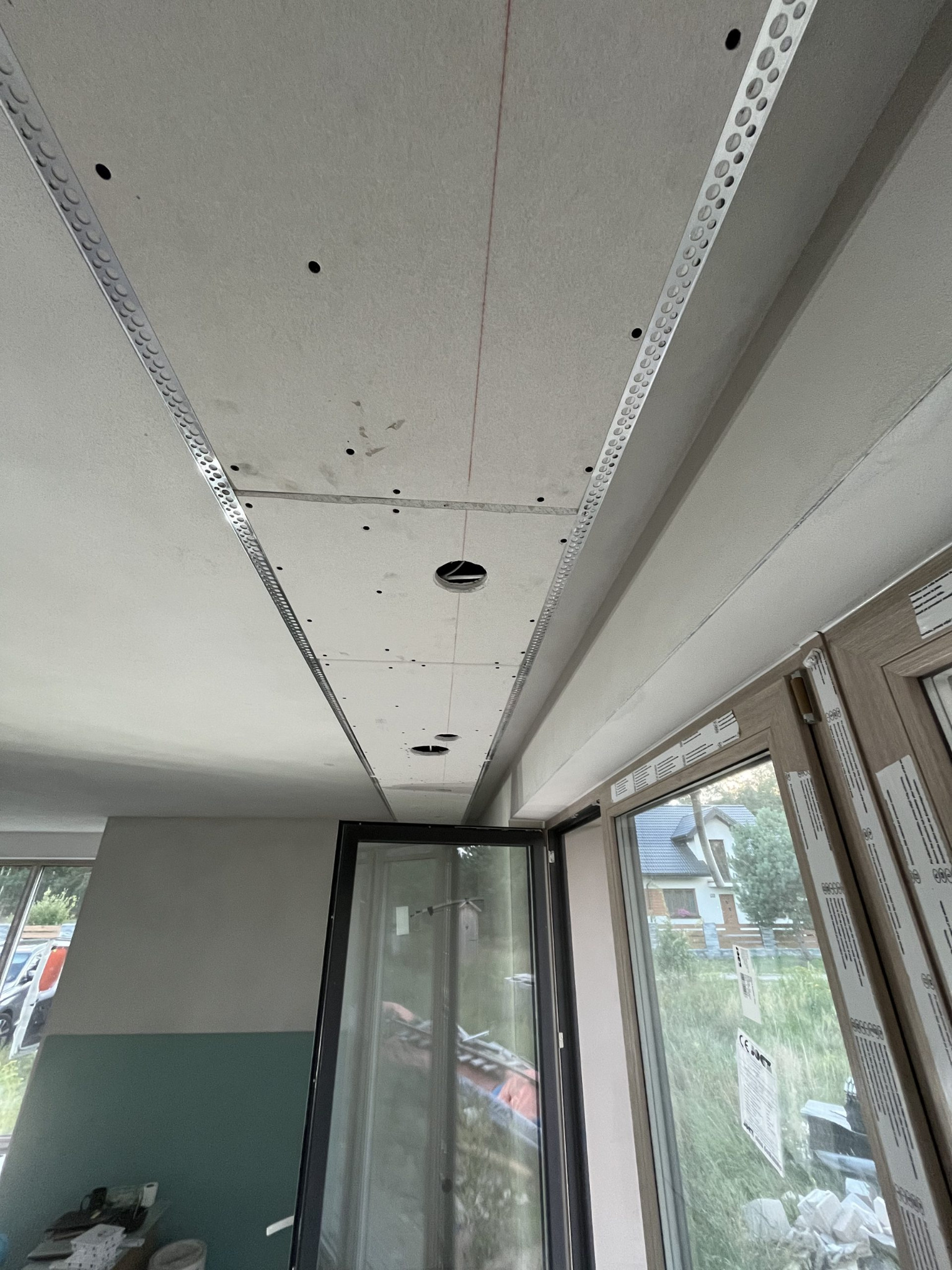 Sufit z zamontowaną płytą karton-gips, widoczne profile i otwory na oświetlenie, zarysowany czerwoną linią. W tle okno z widokiem na dom.