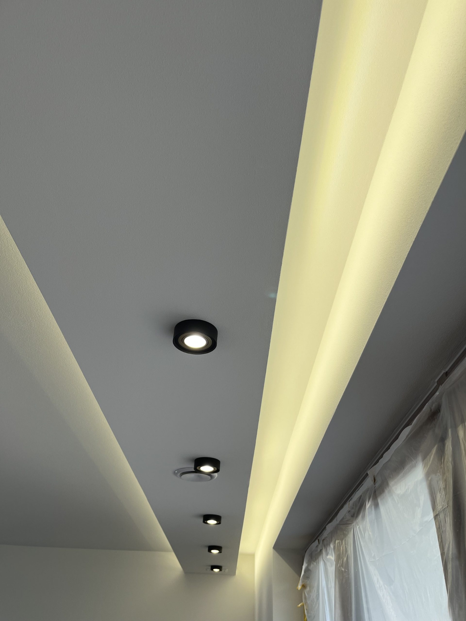 Sufit z oświetleniem LED wzdłuż krawędzi i punktowymi lampami, świeżo pomalowany, z folią ochronną na oknie. Nowoczesne wykończenie wnętrza.