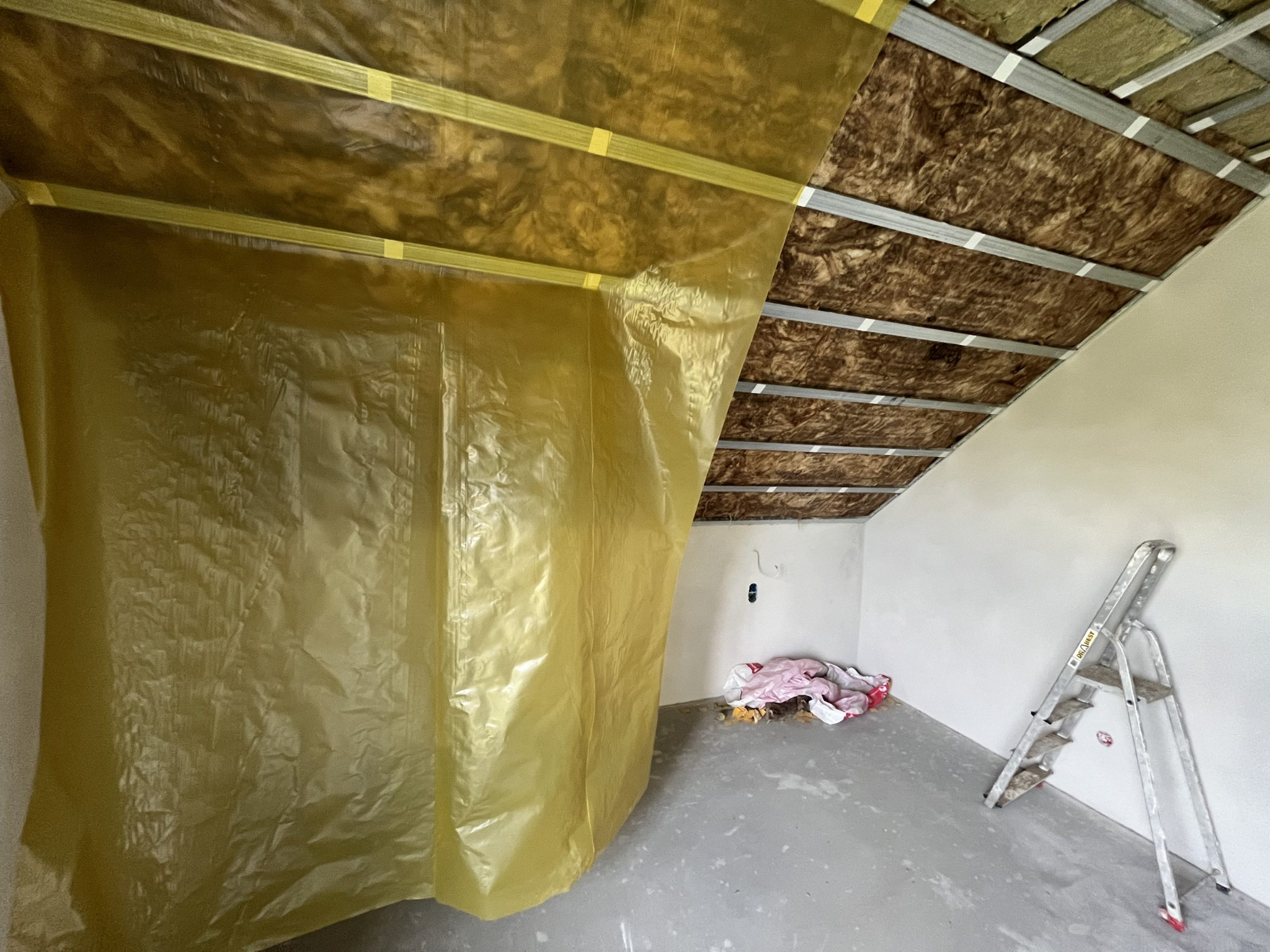 Skośne poddasze w trakcie prac: widoczna izolacja wełną mineralną, metalowy stelaż i folia ochronna. W tle drabina i resztki materiałów budowlanych.